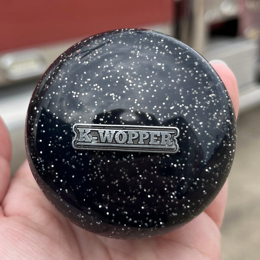 Black speckled knob with 'Wopper' branding held in a hand, blurred vehicle in the background