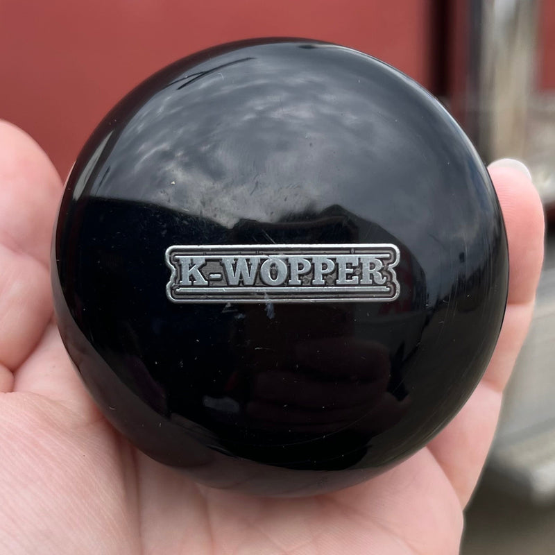Black knob with 'K-Wopper' logo held in a hand, blurred background of a vehicle.