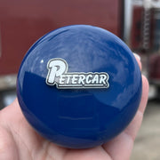 Blue knob with 'PETERCAR' logo held in a hand, with a blurred truck in the background