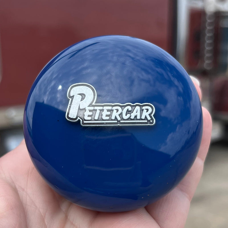 Blue knob with 'PETERCAR' logo held in a hand, with a blurred truck in the background