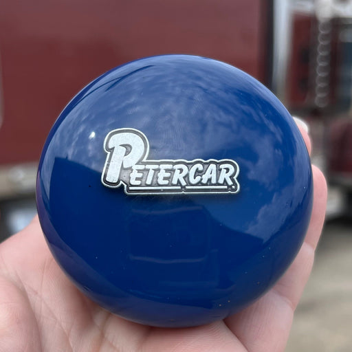 Blue knob with 'PETERCAR' logo held in a hand, with a blurred truck in the background