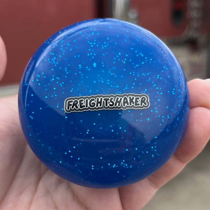 Blue glittery object with 'Freightshaker' logo held in a hand, blurred red vehicle in the background
