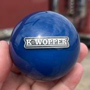 Blue knob with 'K-Wopper' logo held in a hand, blurred truck in the background