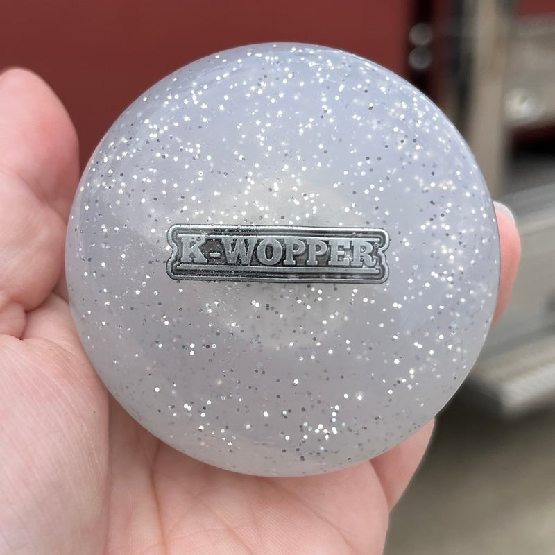 Hand holding a gray glittery ball with 'K-Wopper' branding in front of a red vehicle.