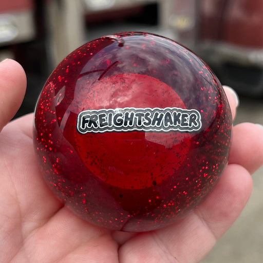 Red glittery ball with 'Freight Shaker' logo held in a hand, with a blurred truck in the background.