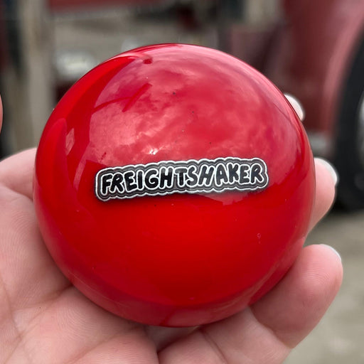 Red ball with 'Freight Shaker' label held in a hand, with a large truck in the background.