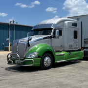 Green and silver semi-truck parked on a concrete lot with a blue sky and clouds in the background.