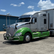 Green and gray semi-truck parked on a concrete surface with a blue sky in the background.
