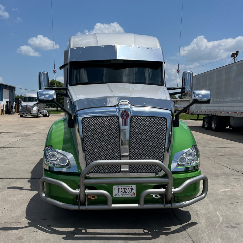 Green and silver semi-truck parked on a concrete surface with a blue sky in the background.
