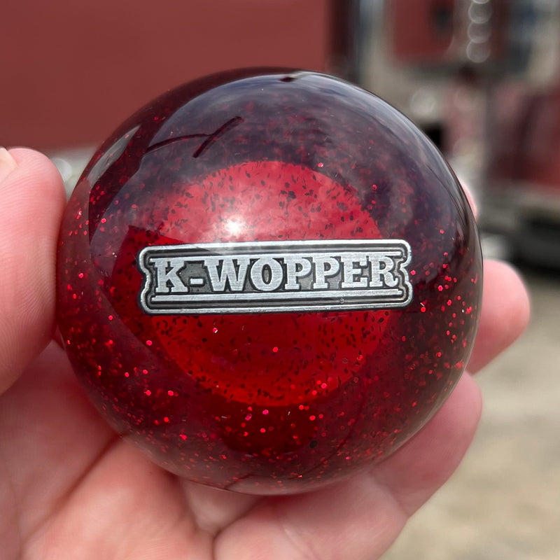 Red knob with 'K-Wopper' logo held in a hand, blurred truck in the background