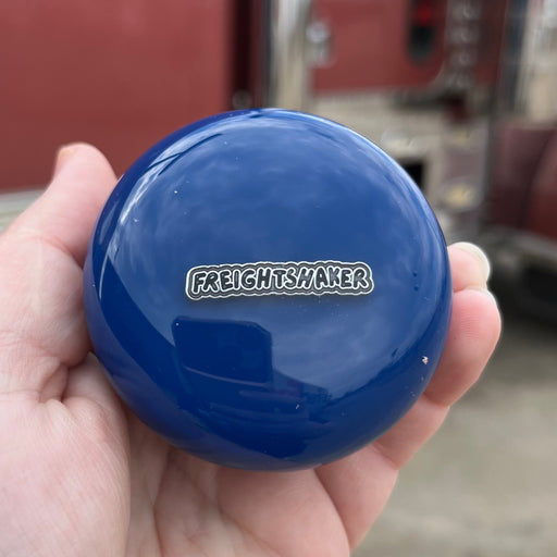 Hand holding a blue round object with 'Freightliner' logo in front of a red truck.