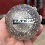 Hand holding a glittery round object with 'K-Wopper' logo in front of a red truck.
