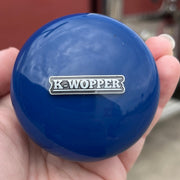 Blue knob with 'K-Wopper' logo held in a hand, blurred red vehicle in the background